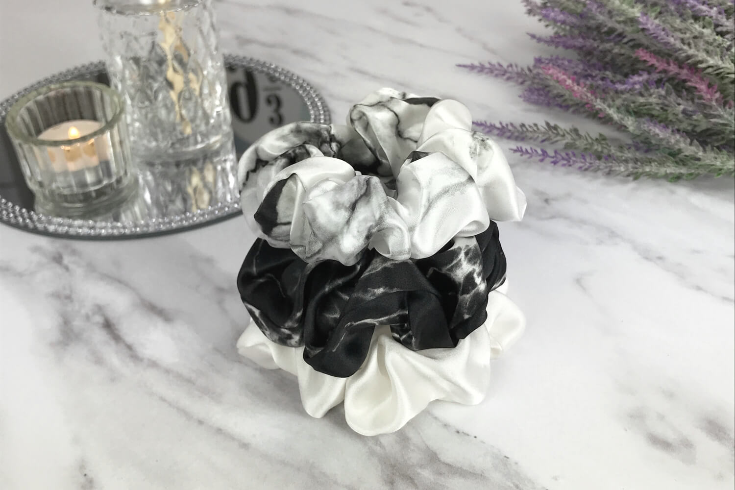 Celestial Silk large white marble, ivory and black marble silk scrunchies stacked on marble counter with lavender plant and a candle in the background