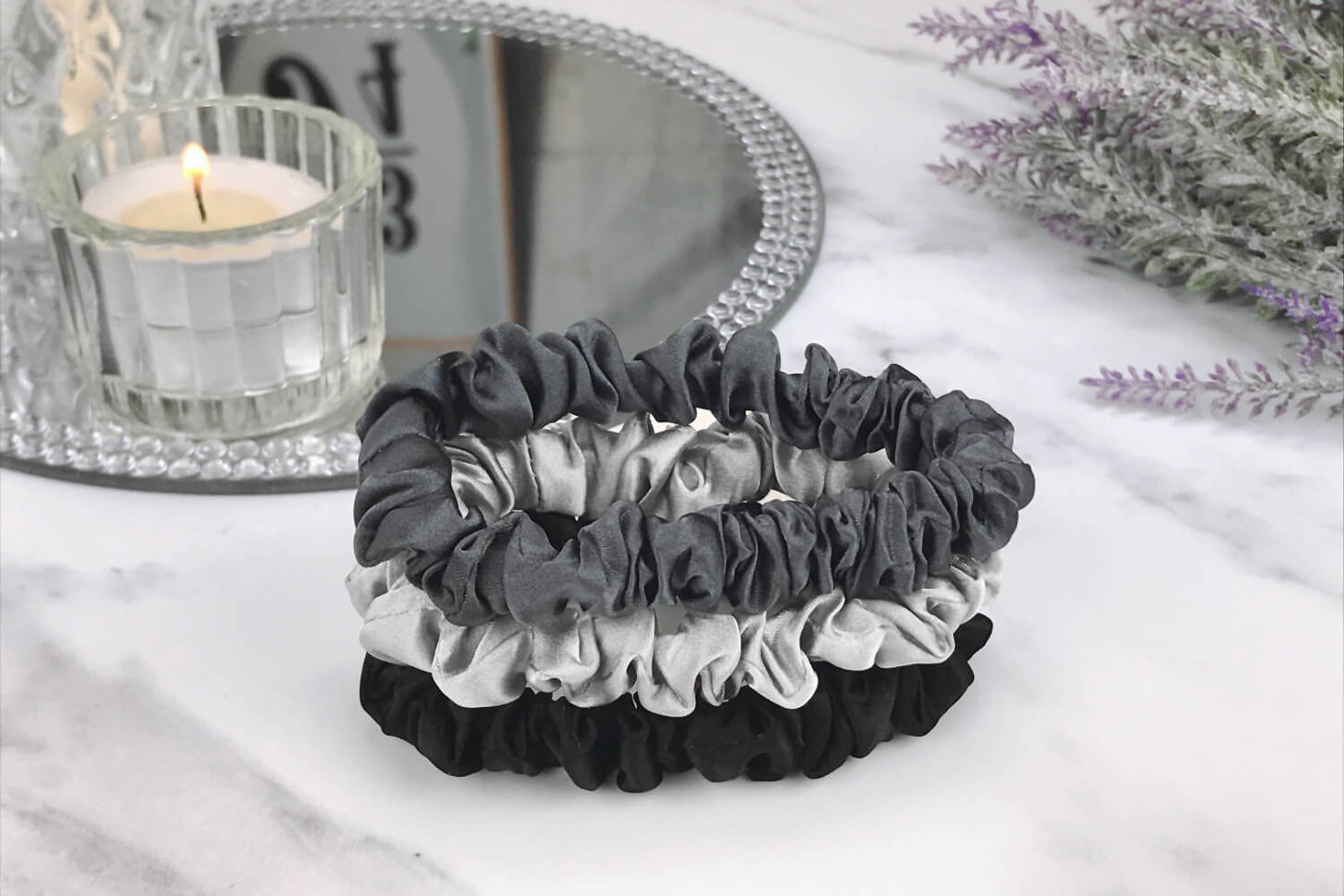 Celestial Silk skinny charcoal black and silver silk scrunchies stacked on marble counter with lavender plant and a candle in the background